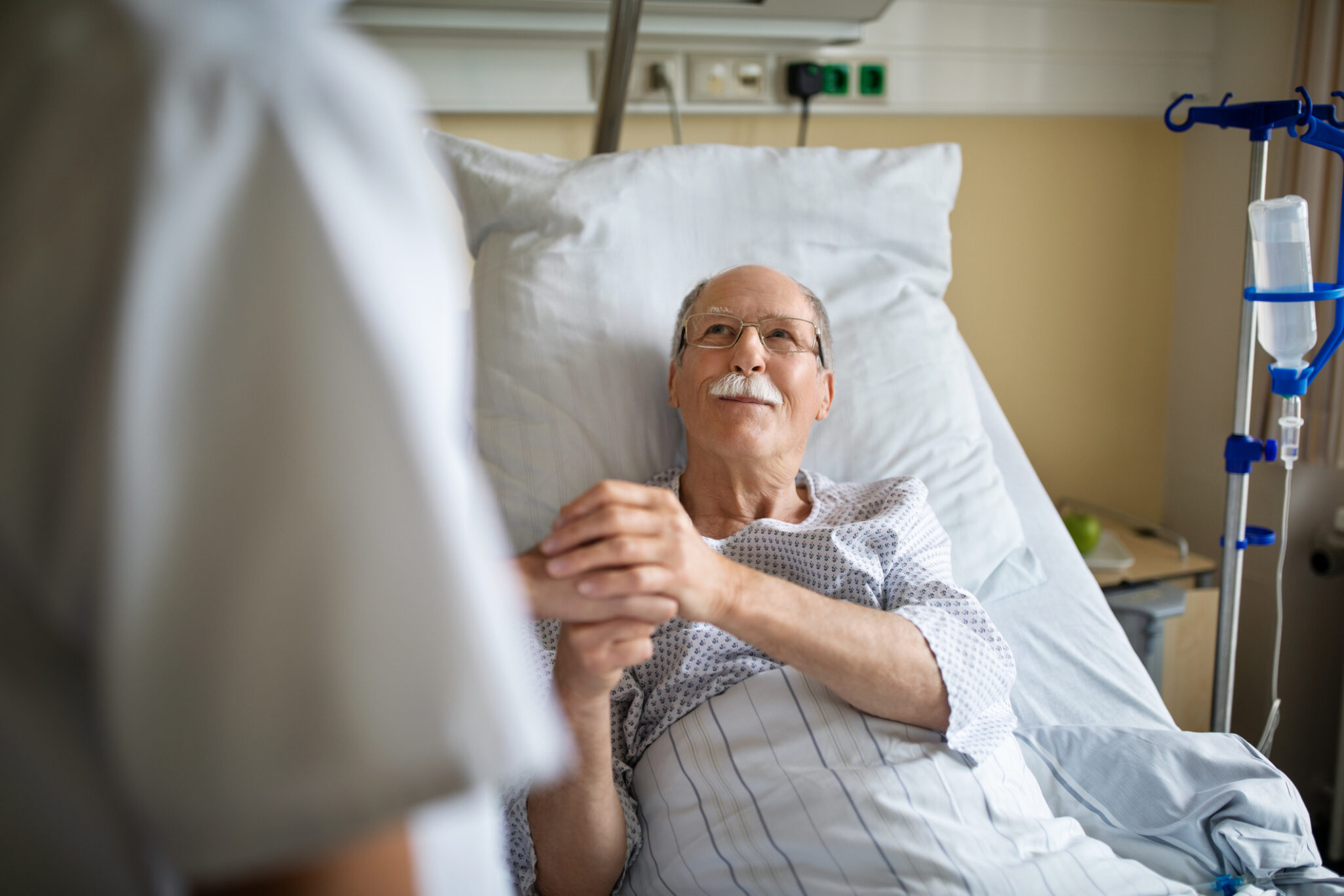 Senior man in a hospital bed holding the hands of a health care professional